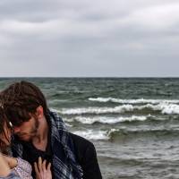Photograph featuring two a male-presenting figure holding a female-presenting one on their arms at a lake shore in intimate interaction